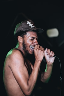 A person wearing a cap and holding a microphone is singing passionately. The person is shirtless, has facial hair, and appears to be sweating. The background is dark, emphasizing the person as the focal point of the image.