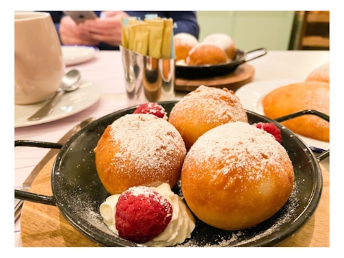 Three golden-brown pastries dusted with powdered sugar are placed in a black skillet, accompanied by fresh raspberries and whipped cream. A cup with a saucer and spoon, sugar packets in a shiny holder, and blurred elements of a dining setting are also visible.