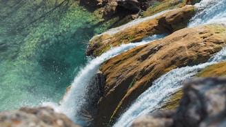 A cascade of clear water flows over layers of smooth, light brown rock. The water appears vibrant and lively as it catches the sunlight, creating a bright, glistening effect. Below, the water in the pool is a vivid turquoise, revealing the rocky bed beneath.