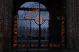 An ornate wrought iron gate featuring elaborate designs that include celestial motifs such as a sun and crescent moon. The gate is set within a brick archway, and the intricate patterns are highlighted by the glow from behind, creating an artistic and mystical ambiance.