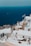 Whitewashed buildings with blue accents are perched on a hillside overlooking a deep blue sea. A Greek flag waves gently in the breeze, and there is a small dog resting in the foreground. A few people can be seen walking along the steps between the buildings.