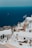 Whitewashed buildings with blue accents are perched on a hillside overlooking a deep blue sea. A Greek flag waves gently in the breeze, and there is a small dog resting in the foreground. A few people can be seen walking along the steps between the buildings.