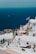 Whitewashed buildings with blue accents are perched on a hillside overlooking a deep blue sea. A Greek flag waves gently in the breeze, and there is a small dog resting in the foreground. A few people can be seen walking along the steps between the buildings.