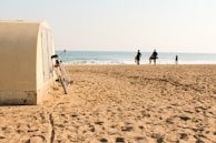 A sandy beach with a small structure on the left side, next to which a bicycle is leaning. In the distance, people are walking towards the ocean carrying surfboards, while others can be seen swimming.