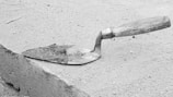 A trowel with a wooden handle and metal blade is resting on a rough concrete or sand surface. The blade is partially covered with a small amount of wet cement.