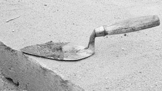 A trowel with a wooden handle and metal blade is resting on a rough concrete or sand surface. The blade is partially covered with a small amount of wet cement.
