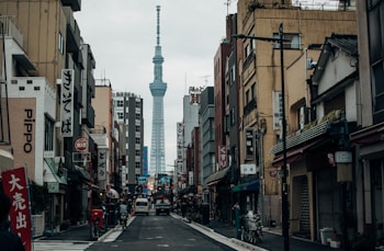 A bustling urban street scene featuring traditional Japanese architecture and various shop signs in kanji, with the Tokyo Skytree towering prominently in the background. The street is lined with buildings on both sides, and a few vehicles and bicycles are present. The atmosphere conveys a typical day in a busy city with muted colors.