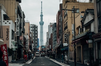 A bustling urban street scene featuring traditional Japanese architecture and various shop signs in kanji, with the Tokyo Skytree towering prominently in the background. The street is lined with buildings on both sides, and a few vehicles and bicycles are present. The atmosphere conveys a typical day in a busy city with muted colors.