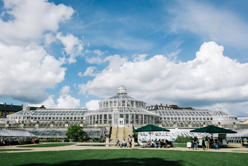 The image features a large, elegant greenhouse with a classical glass architecture, situated in a landscaped area. The greenhouse has multiple domes and wings, giving it a sophisticated and grand appearance. In front of the building, there is a lawn where people are gathered under green umbrellas, adding a lively touch to the scene. The sky is bright with scattered clouds enhancing the overall serenity and beauty of the setting.