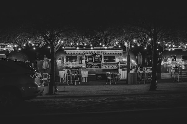 A night scene featuring a retro-style food truck with string lights illuminating the area. Empty chairs and tables are arranged neatly in front of the truck, which has a polished metallic exterior. The setting suggests a quiet street with trees framing the scene, creating a cozy outdoor dining atmosphere.