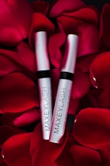 Two silver tubes labeled 'MAXEYLASH eyelash enhancing serum' are nestled amongst vibrant red rose petals.
