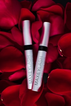 Two silver tubes labeled 'MAXEYLASH eyelash enhancing serum' are nestled amongst vibrant red rose petals.