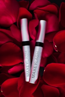 Two silver tubes labeled 'MAXEYLASH eyelash enhancing serum' are nestled amongst vibrant red rose petals.