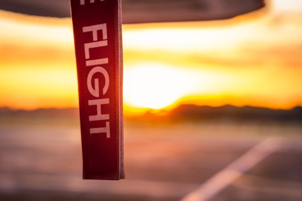 A red tag with white text reading 'FLIGHT', hanging prominently in the foreground with a blurred background of a warm, glowing sunset. The horizon line and abstract shapes suggest an airport setting.