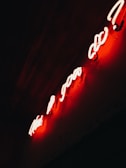 A glowing red neon sign with curved script letters against a dark background creates a dramatic contrast. The neon light casts a warm glow on the surface below, creating an atmospheric and slightly mysterious ambiance.