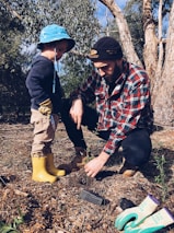 A man and a young child are planting a young tree in a forested area. The man is wearing a plaid shirt and a beanie, while the child is dressed in a blue hat and yellow boots. Gardening gloves and a small trowel are on the ground nearby.