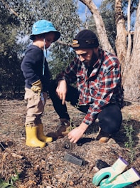 A man and a young child are planting a young tree in a forested area. The man is wearing a plaid shirt and a beanie, while the child is dressed in a blue hat and yellow boots. Gardening gloves and a small trowel are on the ground nearby.