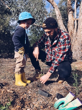 A man and a young child are planting a young tree in a forested area. The man is wearing a plaid shirt and a beanie, while the child is dressed in a blue hat and yellow boots. Gardening gloves and a small trowel are on the ground nearby.