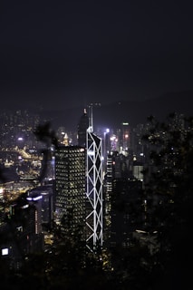 A nighttime cityscape featuring a prominent skyscraper with illuminated geometric patterns. The city lights create a vibrant backdrop against the silhouette of surrounding mountains. The overall scene captures a blend of architecture and natural elements.
