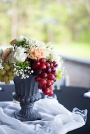 A decorative arrangement featuring a mix of flowers and clusters of grapes in a classic black urn. White, peach, and green flowers are complemented by red and green grapes cascading over the edge. The setting is on a black tablecloth, enhanced by a sheer white fabric draped gracefully around the base of the urn.