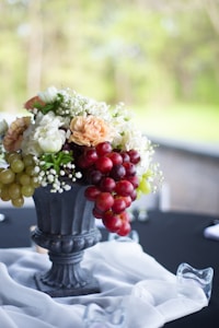 A decorative arrangement featuring a mix of flowers and clusters of grapes in a classic black urn. White, peach, and green flowers are complemented by red and green grapes cascading over the edge. The setting is on a black tablecloth, enhanced by a sheer white fabric draped gracefully around the base of the urn.