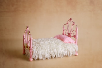 A small, decorative bed with a wrought iron frame painted pink. It features a fluffy white mattress and a single pink pillow. The background is a solid, warm beige color.