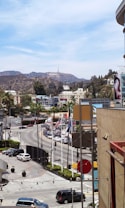 A bustling urban area with cars and palm trees lining the road. In the distance, the Hollywood sign is visible atop the hills, surrounded by scattered buildings and greenery. The foreground includes a busy street scene with flags and traffic.