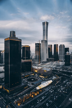 A cityscape featuring several tall, modern skyscrapers mostly in dark tones with golden accents. The tallest building has a sleek, futuristic design. Streets and buildings are illuminated with bright lights, and numerous cars are visible in traffic.