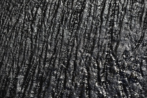 A textured surface with irregular, wavy lines and a glossy appearance. The surface has a dark, almost black color with reflective highlights that give it a slick, shiny look.