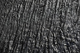 A textured surface with irregular, wavy lines and a glossy appearance. The surface has a dark, almost black color with reflective highlights that give it a slick, shiny look.