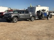 A gray pickup truck is parked on a dirt surface, towing a trailer carrying an off-road vehicle. In the background, there is a white camper and several other vehicles.