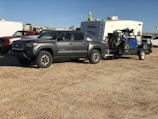 A gray pickup truck is parked on a dirt surface, towing a trailer carrying an off-road vehicle. In the background, there is a white camper and several other vehicles.