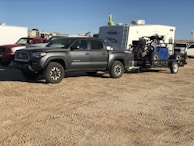 A gray pickup truck is parked on a dirt surface, towing a trailer carrying an off-road vehicle. In the background, there is a white camper and several other vehicles.