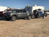 A gray pickup truck is parked on a dirt surface, towing a trailer carrying an off-road vehicle. In the background, there is a white camper and several other vehicles.