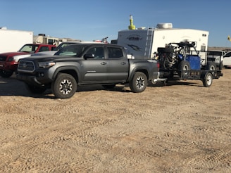 A gray pickup truck is parked on a dirt surface, towing a trailer carrying an off-road vehicle. In the background, there is a white camper and several other vehicles.