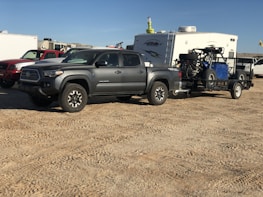 A gray pickup truck is parked on a dirt surface, towing a trailer carrying an off-road vehicle. In the background, there is a white camper and several other vehicles.
