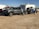 A gray pickup truck is parked on a dirt surface, towing a trailer carrying an off-road vehicle. In the background, there is a white camper and several other vehicles.