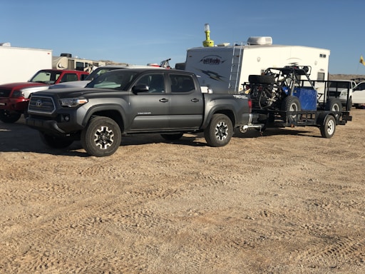 A gray pickup truck is parked on a dirt surface, towing a trailer carrying an off-road vehicle. In the background, there is a white camper and several other vehicles.