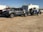 A gray pickup truck is parked on a dirt surface, towing a trailer carrying an off-road vehicle. In the background, there is a white camper and several other vehicles.