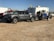 A gray pickup truck is parked on a dirt surface, towing a trailer carrying an off-road vehicle. In the background, there is a white camper and several other vehicles.