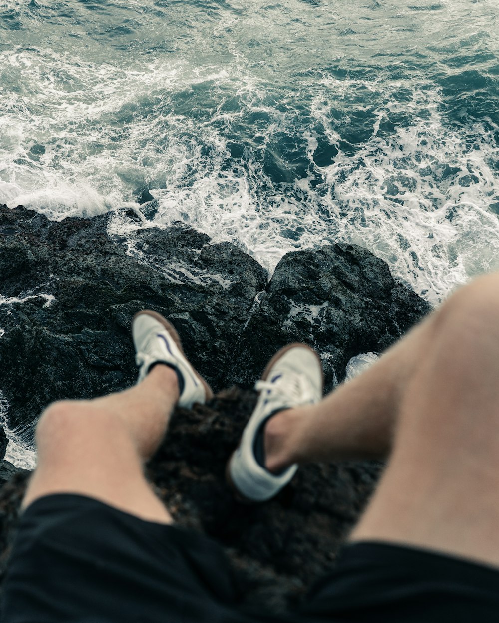 person in black shorts and white sneakers sitting on rock near body of water during daytime