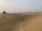 A vast desert landscape with undulating sand dunes stretching into the distance. A white off-road vehicle is parked on one of the dunes, with several people gathered around it. The sky is hazy, and sparse vegetation can be seen scattered across the horizon.