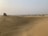 A vast desert landscape with undulating sand dunes stretching into the distance. A white off-road vehicle is parked on one of the dunes, with several people gathered around it. The sky is hazy, and sparse vegetation can be seen scattered across the horizon.
