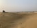 A vast desert landscape with undulating sand dunes stretching into the distance. A white off-road vehicle is parked on one of the dunes, with several people gathered around it. The sky is hazy, and sparse vegetation can be seen scattered across the horizon.