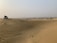 A vast desert landscape with undulating sand dunes stretching into the distance. A white off-road vehicle is parked on one of the dunes, with several people gathered around it. The sky is hazy, and sparse vegetation can be seen scattered across the horizon.