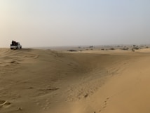 A vast desert landscape with undulating sand dunes stretching into the distance. A white off-road vehicle is parked on one of the dunes, with several people gathered around it. The sky is hazy, and sparse vegetation can be seen scattered across the horizon.