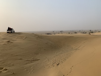 A vast desert landscape with undulating sand dunes stretching into the distance. A white off-road vehicle is parked on one of the dunes, with several people gathered around it. The sky is hazy, and sparse vegetation can be seen scattered across the horizon.