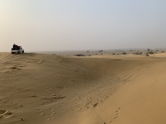 A vast desert landscape with undulating sand dunes stretching into the distance. A white off-road vehicle is parked on one of the dunes, with several people gathered around it. The sky is hazy, and sparse vegetation can be seen scattered across the horizon.
