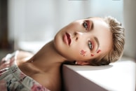 A close-up portrait of a person with artistic makeup, including red and pink eyeshadows and butterfly designs on their cheeks. The individual is gazing directly at the viewer with light-colored eyes. They are lying in a relaxed pose, with their head resting on a white surface. Soft natural light enhances the serene and dreamy atmosphere.