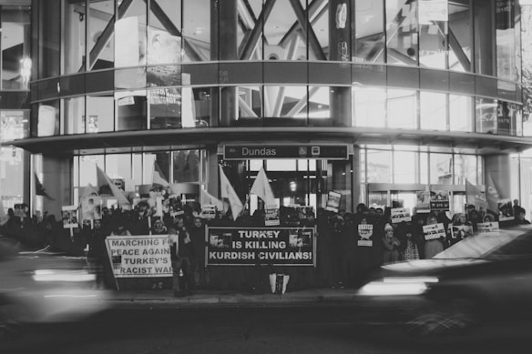 A large group of people are gathered outside a building, holding banners and signs with messages protesting against Turkish military actions. The scene captures a moment of activism and demonstration, with some individuals holding flags. It's a nighttime setting with vehicles appearing in motion in the foreground.